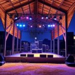 Palco de evento musical Quinta do Pop e Rock em Águas Claras, com equipamentos de som e luzes, representando estreia na cena musical brasileira.