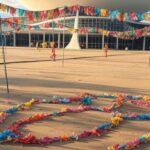 Banners de eventos de carnaval antecipado em praça de Brasília, com arquitetura icônica ao fundo, criticando diluição de tradições.