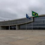 Fachada da CLDF em Brasília, contrastando com moradias precárias ao fundo, simbolizando ignorância a urgências sociais em sessão solene para corretores de seguros.