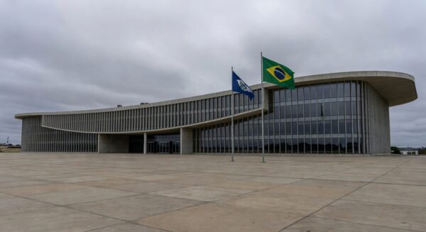 Fachada da CLDF em Brasília, contrastando com moradias precárias ao fundo, simbolizando ignorância a urgências sociais em sessão solene para corretores de seguros.
