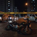 Cena de crime em rua de Taguatinga com fita de isolamento, motocicleta caída e viaturas da Polícia Militar após ataque a tiros.